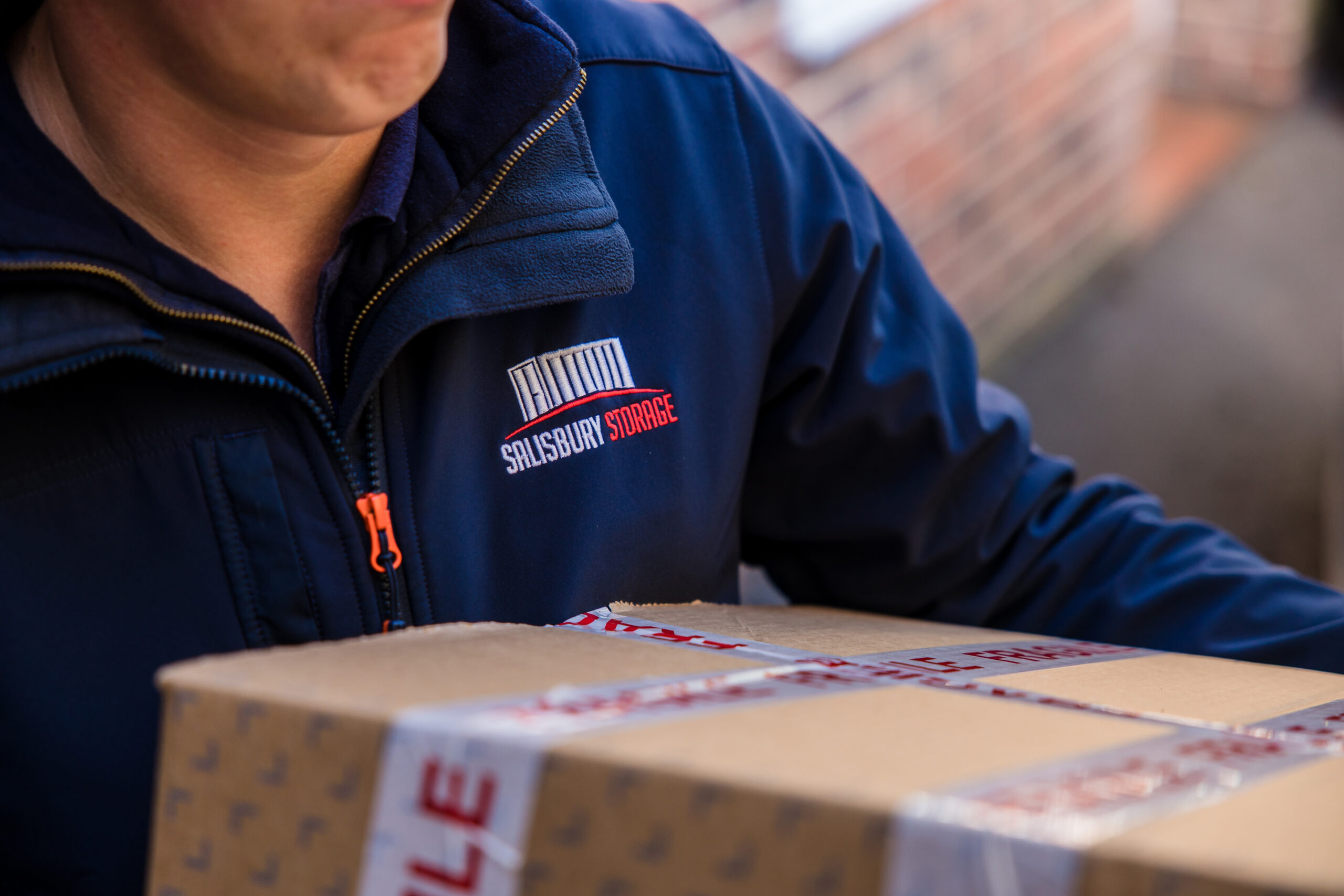A man in a dark blue salisbury storage jacket carrying a box outside.