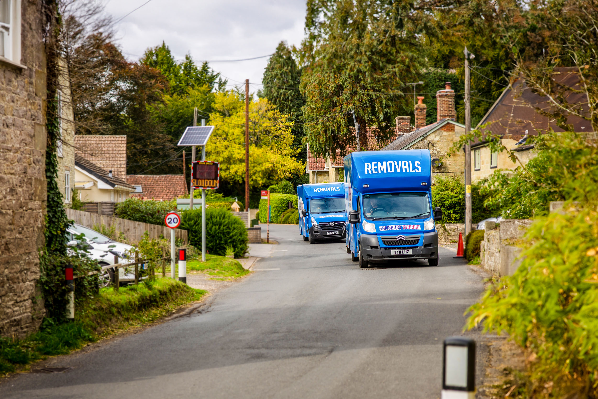 Two blue removals and storage vans driving down the road in beautiful surroundings.