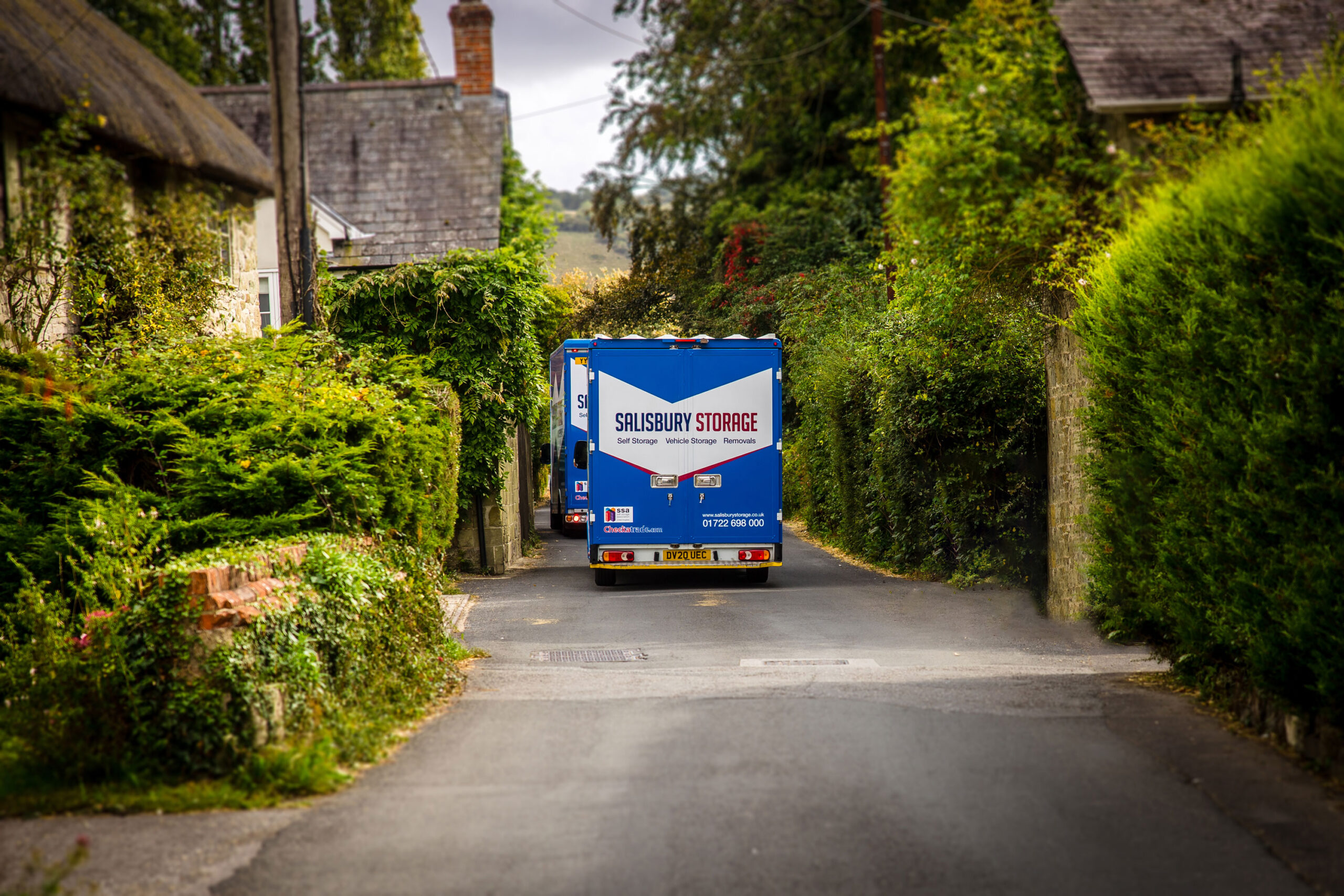Two blue removals and storage vans driving into the distance on a village road, lots of green scenery.