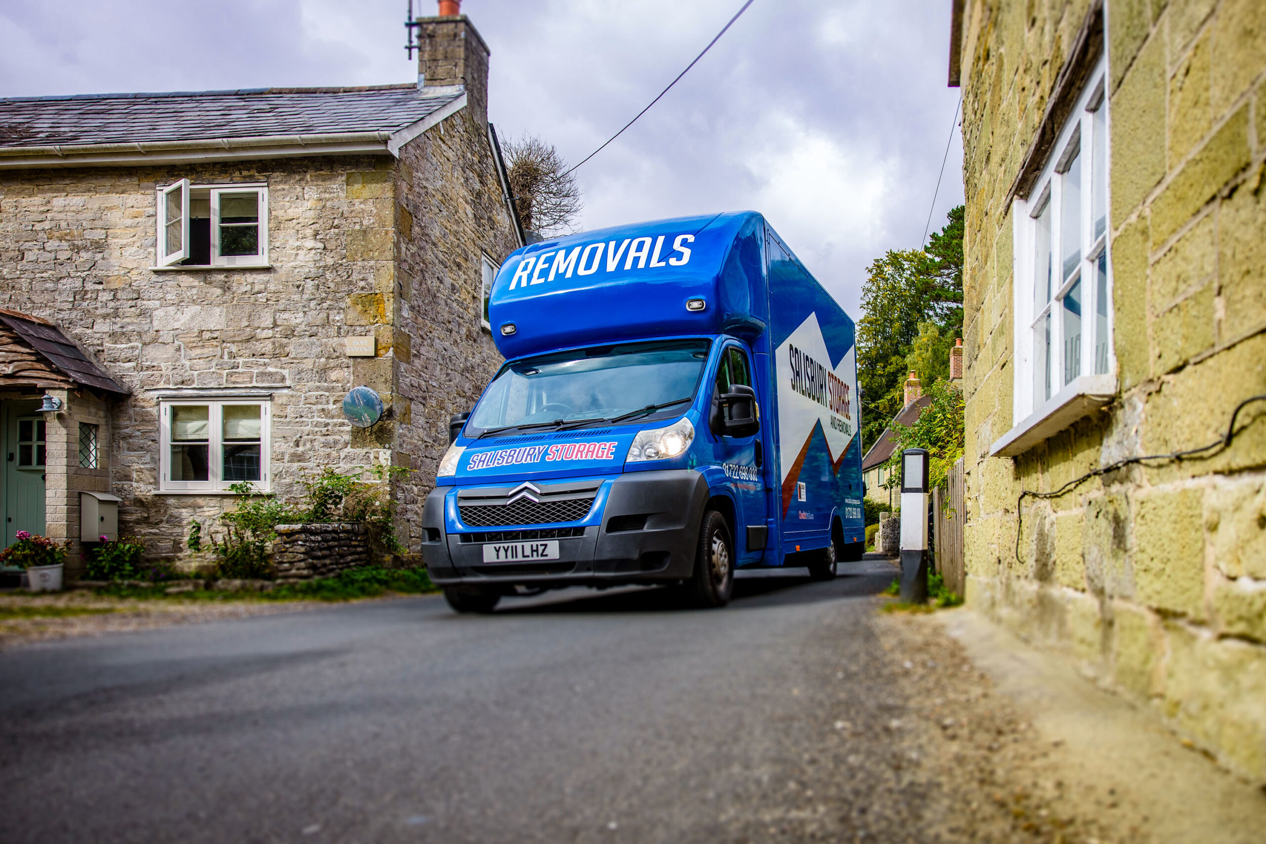 A blue removals van driving down a winding village road, surrounded by greenery and buildings.